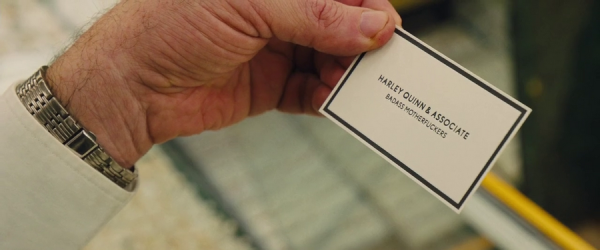 A man's left hand holds a business card, white with black border and text, that reads, in block capitals, 'Harley Quinn & Associate. Badass motherfuckers'.