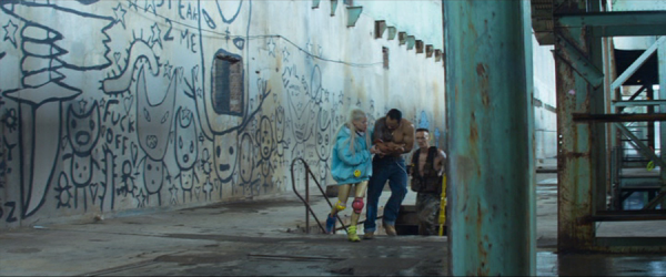 Scene from a derelict city area shows three characters hurrying up from underground steps, one of them visibly injured. The high wall to the left is filled with crude graffiti of cartoonish stick-aliens and some text, including 'Fuck off'.