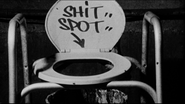 Front view of a makeshift toilet, with open seat balanced on some poles over a bucket. The inside lid has text that says, in all capitals, 'Shit spot' and an arrow pointing down.
